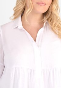 Biała bawełniana bluzka z kołnierzem na zgięciu, z zapięciem na guziki oraz marszczonym materiałem poniżej biustu. Lekka i lekko przezroczysta tekstura.