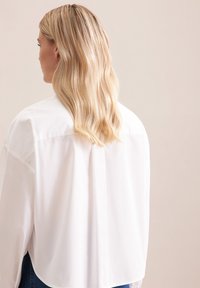 Camicia bianca oversize con piega sul retro, orlo curvo e spalle senza cuciture. Tessuto fluido, texture liscia, abbinata a onde morbide.