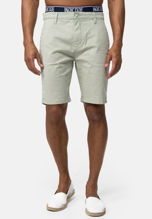 Lichtgroene gebloemde shorts met een knoop en rits, voorzien van een marineblauwe tailleband met "INDICODE" branding. G gedragen met witte espadrilles.