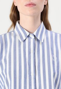 Blauw-wit gestreepte shirt met een knoopsluiting kraag, zachte stof en een geborduurd logo detail aan de linkerkant.