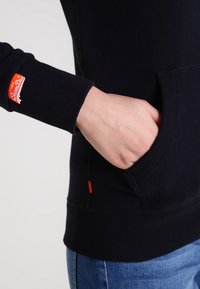 Svart sweatshirt med ribbad mudd, med en orange märkeslapp. Inkluderar en framficka och en slät textur. Sedd från sidan.