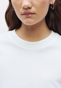 Gaiši pelēks džemperis ar apaļas formas kakla izgriezumu. Gluda tekstūra, minimālistisks dizains, ar smalku ribotu apkakles apdari. Redzamas sudraba stīpas auskari.