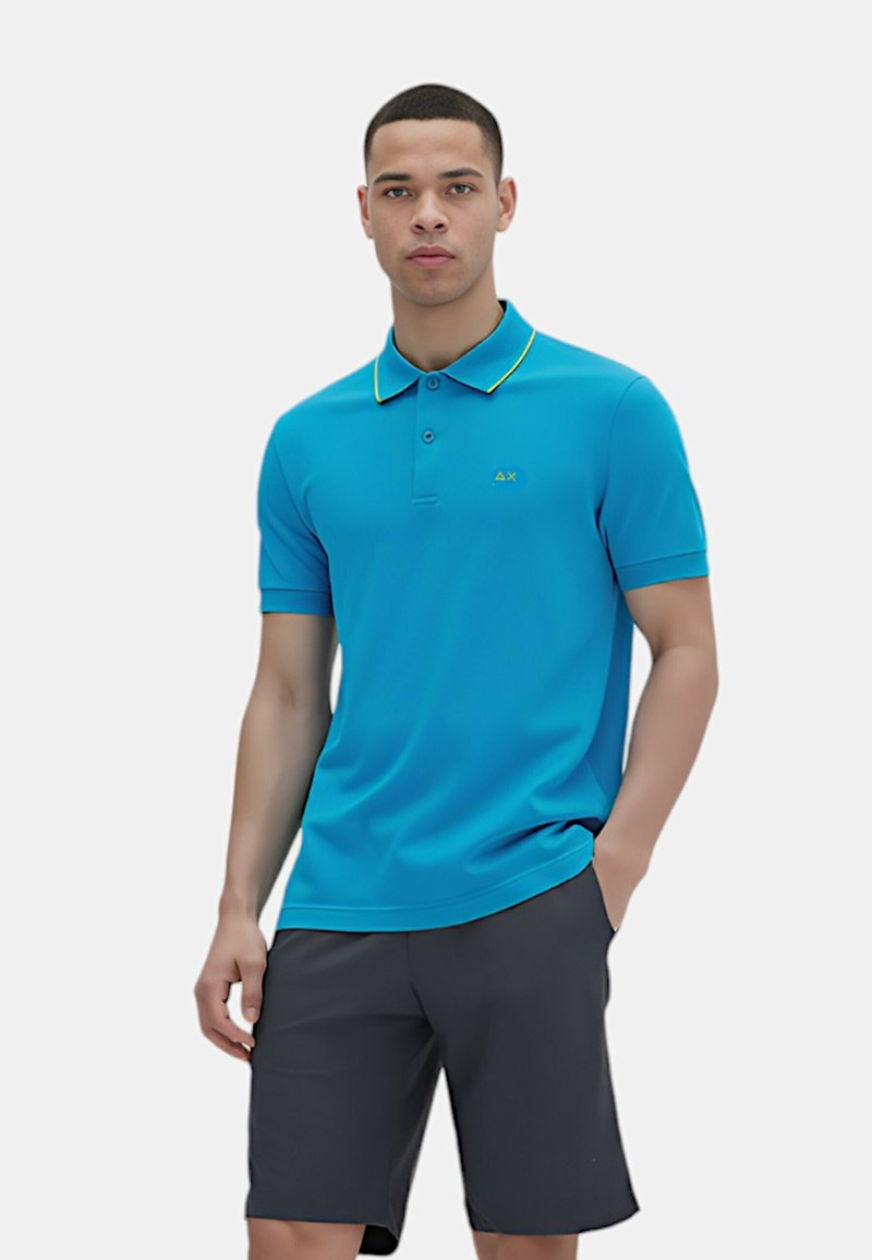 Polo blu realizzato in tessuto liscio, con colletto a righe gialle e un piccolo logo sul petto, abbinato a pantaloni corti scuri.