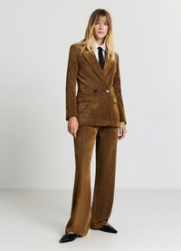 Costume en velours côtelé marron avec blazer croisé et pantalons à jambes larges. Comprend un revers cranté et deux boutons, associé à une chemise blanche et une cravate noire.