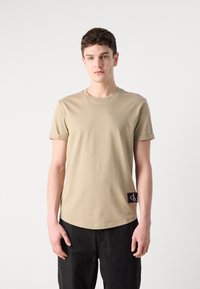 Tricou bej cu mânecă scurtă din bumbac moale, având un guler rotund și un insignă neagră pe partea stângă.