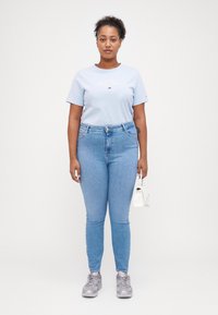 T-shirt en coton bleu clair à manches courtes avec logo, associé à un jean bleu clair taille haute et des baskets grises. Sac à main blanc en accent.