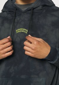 Tmavě šedá mikina s batikovaným vzorem a klokankou, na prsou s vyšívaným žlutým текстem "GOOD FOR NOTHING", se stahovacími tkaničkami.