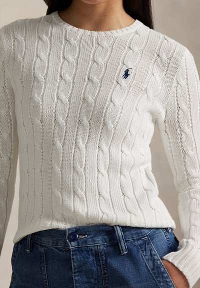 Witte kabelgebreide trui met lange mouwen en marineblauw geborduurd logo, gedragen en ingestopt in een blauwe spijkerbroek.