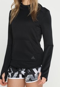 Donna che indossa una felpa nera a maniche lunghe con fori per i pollici e pantaloni corti con motivo bianco e nero, in piedi con le mani vicino ai fianchi.