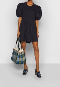Black tiered dress with puff sleeves, paired with a patterned tote bag in blue, green, and cream. Black heeled loafers complete the outfit.