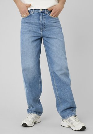 Personne portant un jean droit bleu clair, des baskets blanches et une chemise blanche avec les mains dans les poches sur un fond uni.