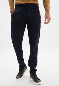 Granatowe joggery z miękkiego materiału, z brązowymi akcentami przy kieszeniach, elastycznym paskiem w talii ze sznurkiem i zwężającym się krojem nogawki.