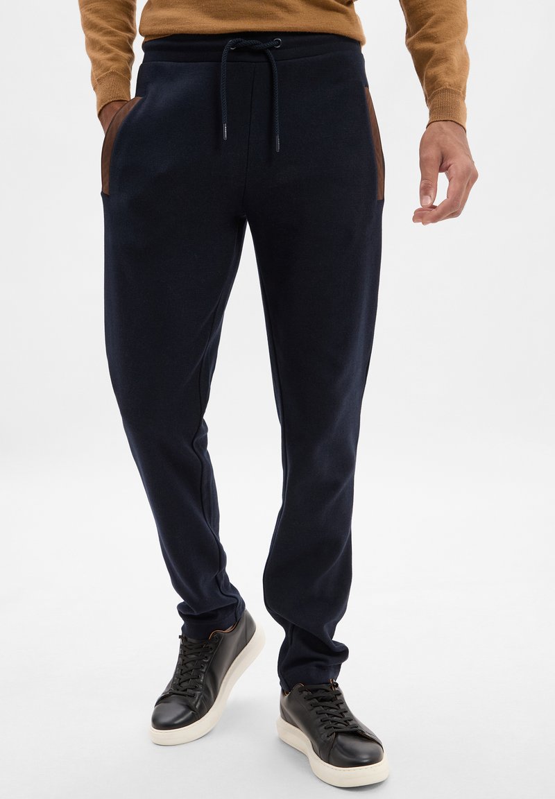 Granatowe joggery z miękkiego materiału, z brązowymi akcentami przy kieszeniach, elastycznym paskiem w talii ze sznurkiem i zwężającym się krojem nogawki.