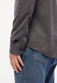 Mörkgrå långärmad skjorta med knäppta manschetter, ihop med ljusblå jeans. Tyget verkar mjukt med en lätt textur.