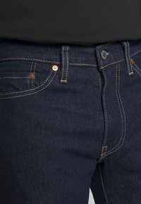 Gros plan sur un jean en denim bleu foncé avec des coutures visibles, des passants de ceinture, des rivets métalliques et une fermeture à bouton, porté avec un haut noir.