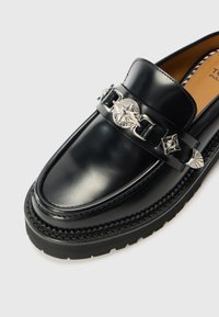 Black leather loafer with a glossy finish, featuring a decorative silver chain and emblem. Thick, ridged black rubber sole with intricate stitching.