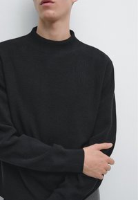 Jeune personne portant un pull noir côtelé, les bras croisés, sur un fond blanc uni, tête partiellement hors cadre.