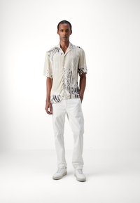 Short-sleeve beige shirt with black abstract print, paired with white pants and white sneakers. Material appears lightweight and smooth.