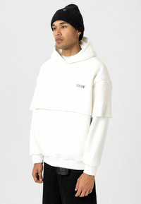 Witte oversized hoodie met een gelaagd ontwerp en korte mouwen, voorzien van een klein logo aan de voorkant. Gecombineerd met zwarte broek en een beanie.