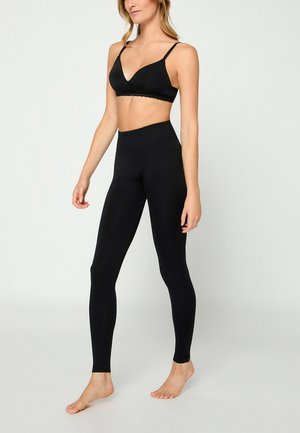 Frau in schwarzem Sport-BH und hoch taillierten schwarzen Leggings, barfuß vor einem einfarbigen hellen Hintergrund stehend.