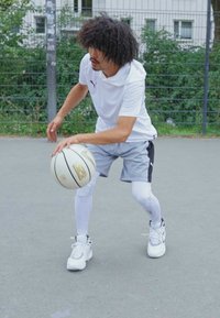 Un giocatore di basket in una maglietta atletica bianca e pantaloni corti grigi dribbla un pallone su un campo. Indossa scarpe da ginnastica bianche e lunghe maniche bianche per le gambe.