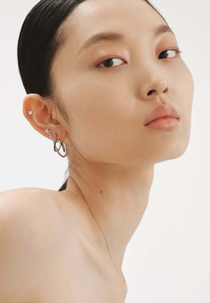 Pendientes de aro colgantes en plata con un acabado texturizado, combinados con pequeños pendientes de botón. La piel del modelo es suave, con un fondo neutro.