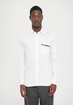 Chemise blanche à manches longues avec boutonnières, dotée d'une poche poitrine avec une étiquette noire. Tissu lisse avec un col classique et un léger éclat.