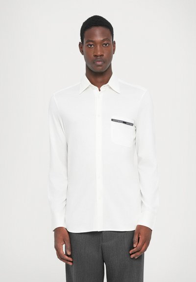 Chemise blanche à manches longues avec boutonnières, dotée d'une poche poitrine avec une étiquette noire. Tissu lisse avec un col classique et un léger éclat.