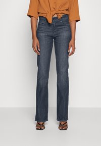 Jeans acampanados de mezclilla azul oscuro con una textura suave, cintura media y diseño de cinco bolsillos, combinados con una blusa color óxido anudada.