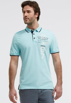 Mann mit braunen Haaren, der ein hellblaues Poloshirt mit marineblauem Besatz und dem Schriftzug "Antigua Fleet" sowie graue Hosen trägt, steht vor weißem Hintergrund.