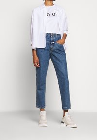 Camisa blanca de botones sobre una camiseta gráfica blanca, combinada con jeans cortos azules y zapatillas beige con acentos texturizados.