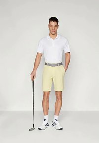 Polo branco, calções amarelos, cinto cinzento. Homem de pé com taco de golfe, usando meias brancas e sapatilhas de ténis. Materiais suaves.