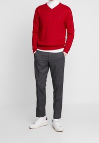 Mies, joka on pukeutunut punaiseen V-pääntiepaitaan, valkoiseen turtleneck-paitaan, harmaisiin muotihousuihin, valkoisiin sukkiin ja valkoisiin lenkkkenkiin, seisoo kädet taskuissa.