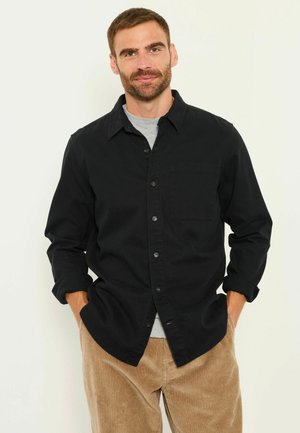 Man with short brown hair and beard wearing a black button-up shirt over a gray t-shirt and tan corduroy pants, hands in pockets.