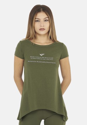 Giovane donna che indossa una maglietta verde oliva con testo positivo sulla bellezza, la positività del corpo, la sostenibilità e il potere delle donne.