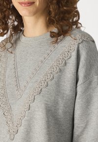Sweatshirt gris avec un col en V orné d'une bordure en dentelle gris clair le long des épaules et sur le devant. Texture douce.