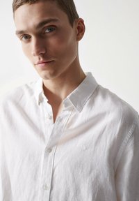 Camisa blanca de lino con botones y cuello, que presenta una sutil textura, un pequeño logo en el pecho y un corte relajado.