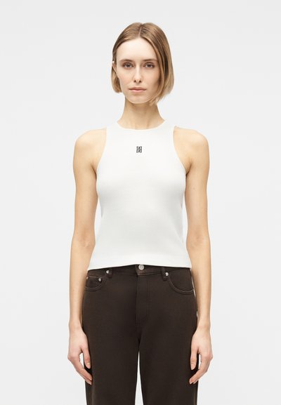 Femme aux cheveux blonds courts portant un haut blanc sans manches côtelé avec un petit logo noir et un pantalon taille haute marron foncé.