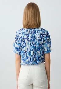 Blusa blu a fantasia con maniche corte, caratterizzata da un mix di forme blu chiare e scure su un tessuto trasparente. Indossata con pantaloni bianchi a vita alta.
