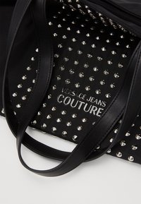 Černá taška přes rameno se zdobeným designem, s kovovými prvky v barvě stříbra, koženými rukojeťmi a logem "VERSACE JEANS COUTURE" na strukturovaném povrchu.