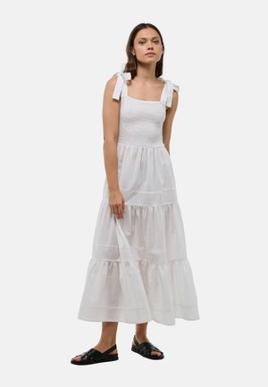 Femme aux longs cheveux bruns portant une robe blanche sans manches avec des nœuds sur les épaules et des sandales noires à bout ouvert, debout devant un fond blanc.