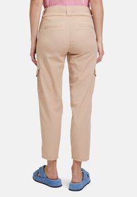 Beige Cargo-Hosen mit Seitentaschen, einem figurbetonten Bund und schmalen Beinen. Getragen mit blauen Schlappen.