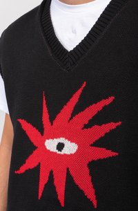 Svart strikket vest med en rød piggete design og et sentralt hvitt øye. V-hals med teksturert ribbing. Bæres over en hvit skjorte.