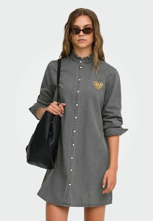 Mujer con vestido gris abotonado con parche de corazón con estampado de leopardo, gafas de sol y lleva un bolso de hombro negro sobre fondo blanco.