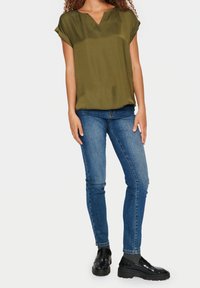 Olijfgroene blouse met korte mouwen en een v-hals, voorzien van een subtiel stippenpatroon, gecombineerd met blauwe skinny jeans en zwarte instappers.