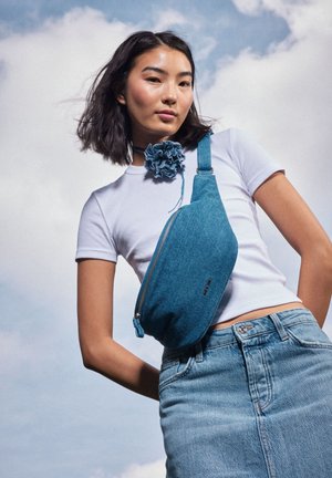 Borsa in denim turchese con chiusura a zip e tracolla regolabile, indossata sopra una camicia bianca aderente e una gonna in denim abbinata.