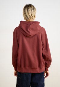 Bordo kapuce ar strukturētu, ribotu dizainu, kas izceļas ar lielu kapuci un elastīgiem piedurknēm. Aizmugurē ir brīvs griezums un nolaižamās plecu daļas.