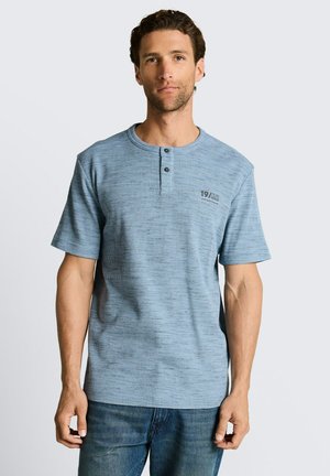 Mann trägt ein blaues Henley-Shirt mit kurzen Ärmeln, zwei Knöpfen und der Aufschrift "19/62" auf der Brust, kombiniert mit blauen Jeans, steht vor einem schlichten Hintergrund.