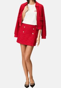 Veste rouge texturée avec fermeture éclair, associée à une mini-jupe rouge ajustée ornée de boutons dorés, et chaussures noires à bout pointu.