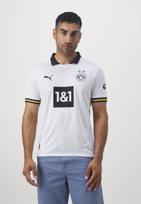 Bela nogometna majica z črnimi ovratniki, z rumenimi in črnimi proženimi rokavi, logotipom z zvezdami in črnim kvadratom s tiskom "1&1".